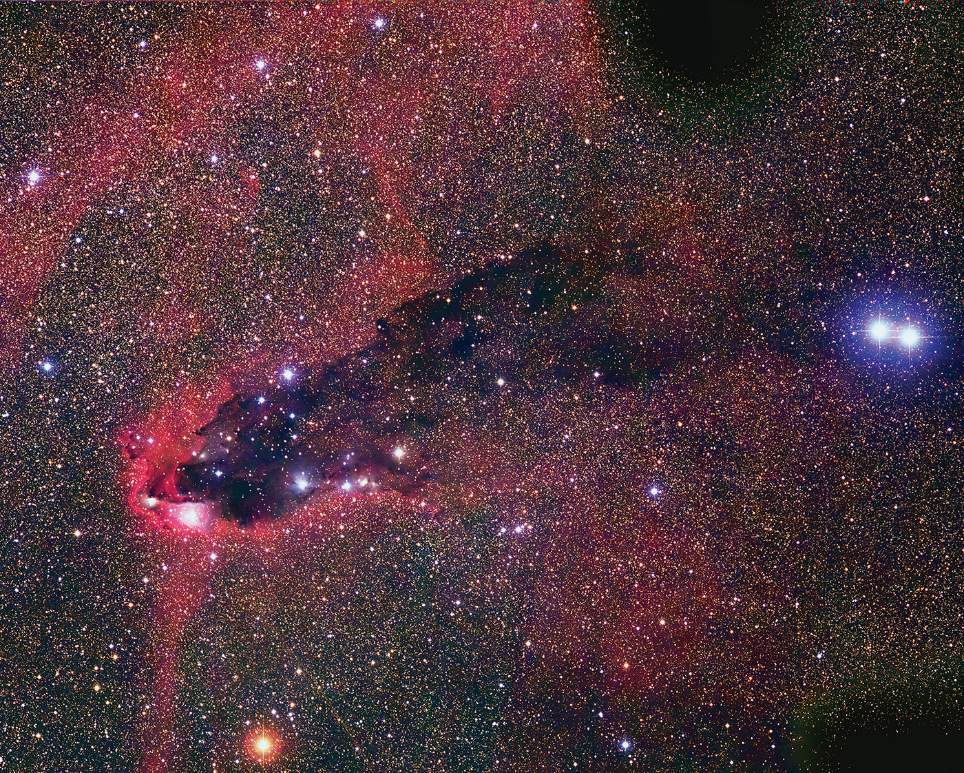 
Denne snodige samlingen av ikke navngitte lyse og mørke tåker er massiv. Den bakoversvøpte formen skyldes stråling fra svært lyssterke stjerner (spesielt Scorpii zeta-1) i den åpne stjernehopen NGC 6231 (ikke i bildet), Scorpius OB-assosiasjonen med svært varme, unge stjerner. Hvis den mørke tåken i bildet er i samme avstand fra Solen som NGC 6321 (omkring 6000 lysår), så er den åpne hopen og den mørke skyen den fordamper minst 100 lysår fra hverandre. Retningen mot strålingskilden ses fra strømningsmønsteret i den mørke skyen og de utpregede, røde kantene (emisjonståker) hvor gassrikt støv utsettes for strålingen fra de varme stjernene i NGC 6321.Lambda Print (Bilde og tekst (editert) © 2000-2010, Australian Astronomical Observatory/David Malin Images)